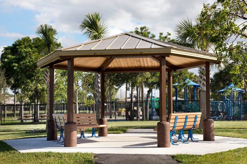 Park gazebo with bench seating surrounded by palm trees and playground in the background.