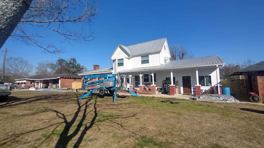 Two-story white house with a metal roof and blue lift in the front yard under a clear blue sky.