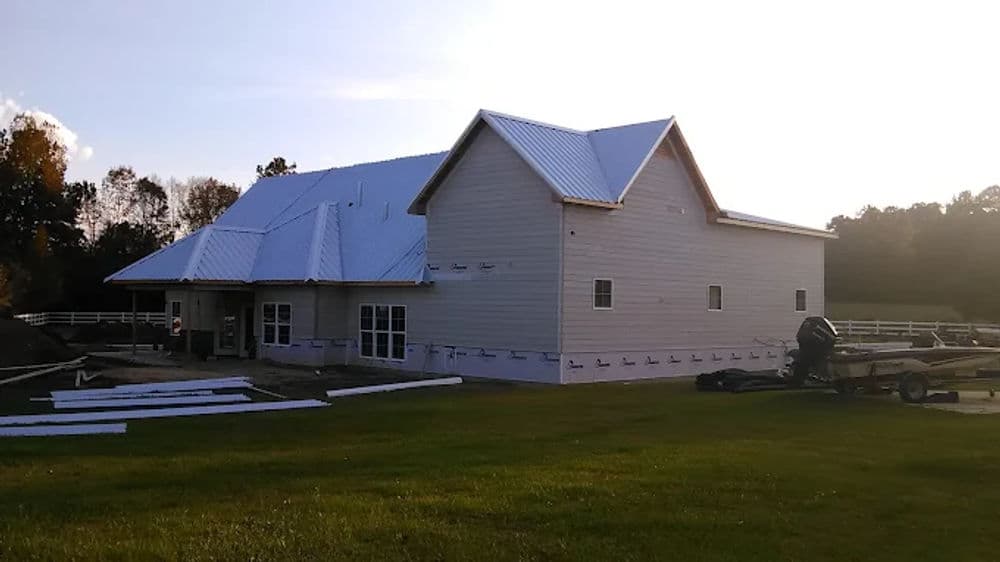 Modern farmhouse with a metal roof under construction, surrounded by green lawn and trees.