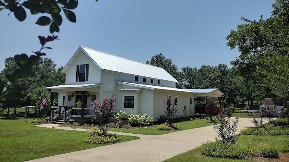 White farmhouse with a metal roof surrounded by green landscape and colorful flowerbeds.