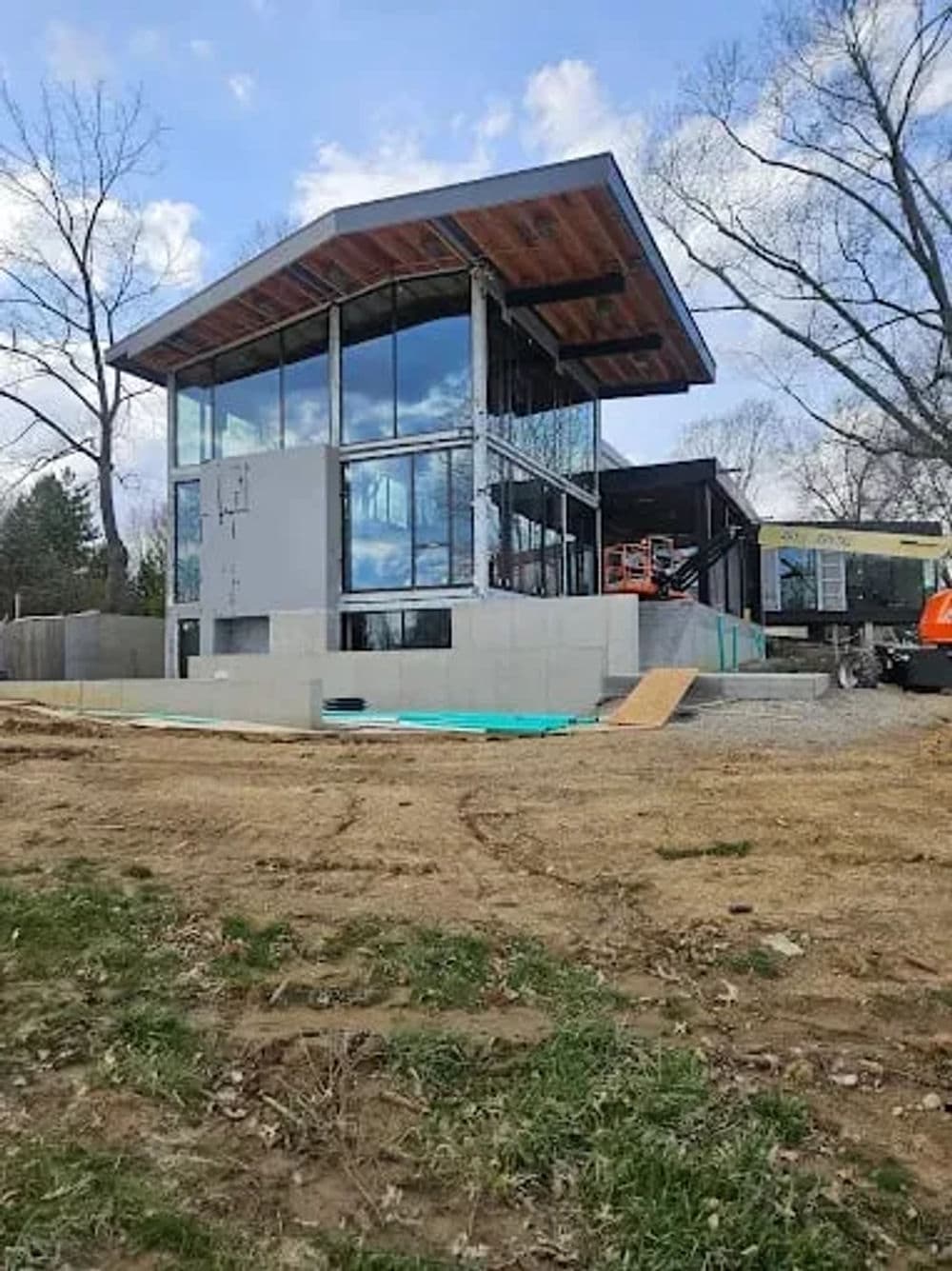 Modern glass-walled house under construction with a wooden roof and surrounding landscaping.