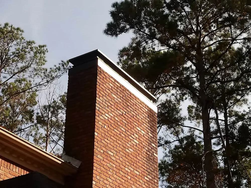 Chimney on a brick house surrounded by tall trees and a blue sky.