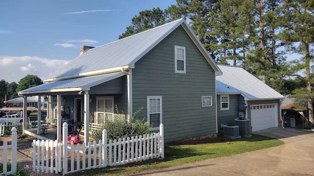 Gray two-story house with white picket fence and modern metal roof. Surrounded by trees.