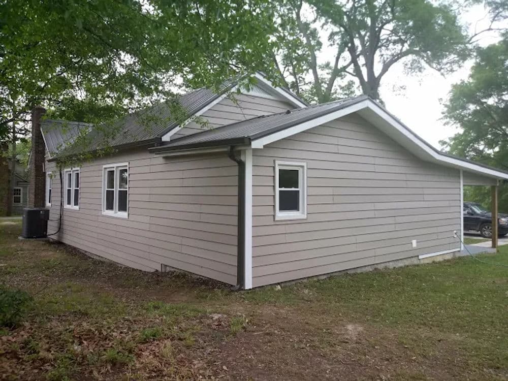 Side view of a modern single-story home with gray siding and large windows, surrounded by greenery.