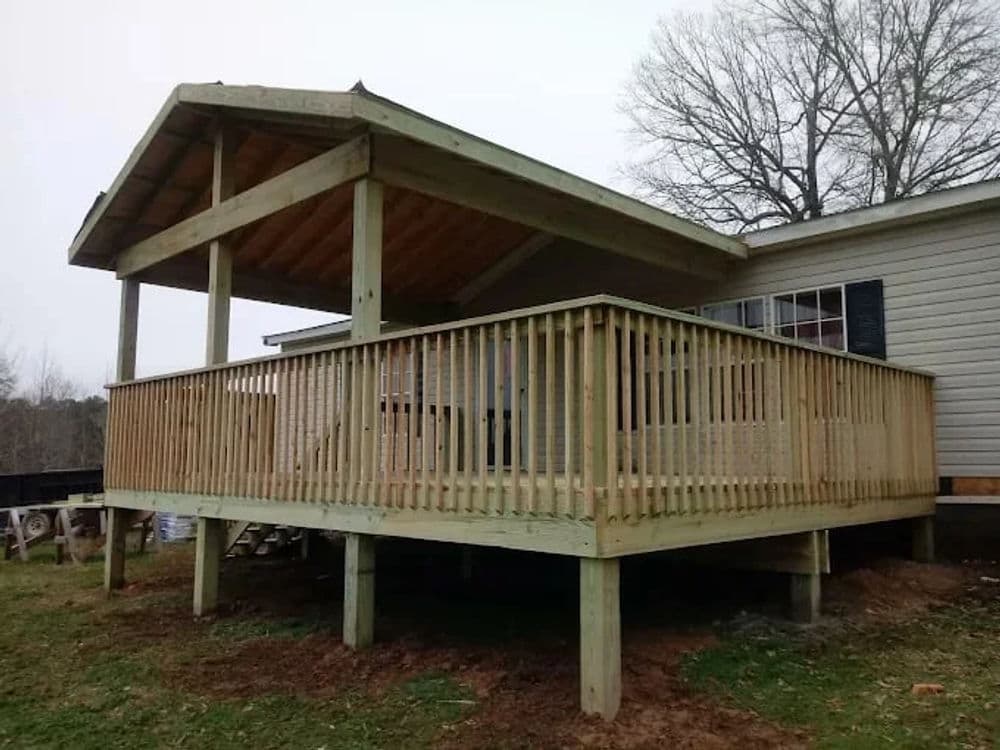 Wooden deck with a covered porch attached to a house, surrounded by grass and trees.