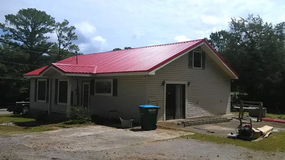 One-story house with red metal roof, green trash bin, and construction tools outside.