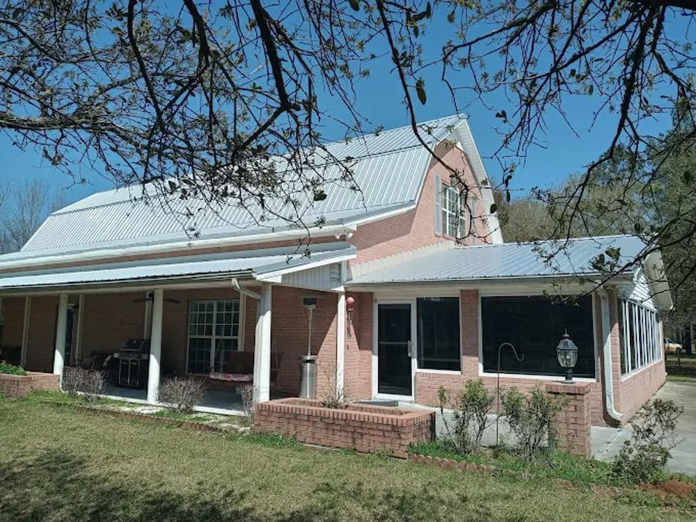 Two-story brick house with a metal roof and porch, surrounded by greenery and trees.