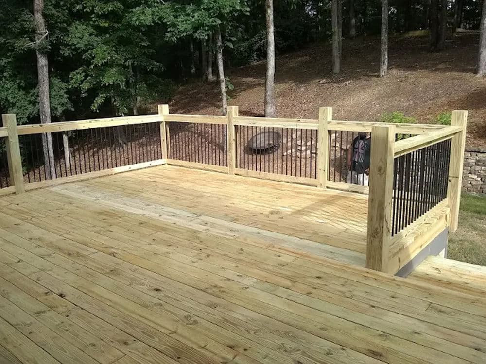 Wooden deck with black railing surrounded by trees and a firepit in the background.