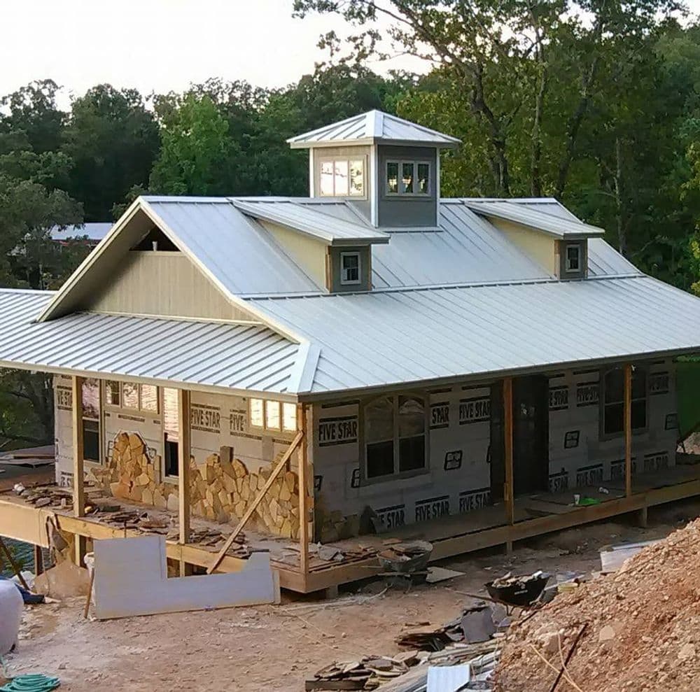 Modern house under construction with a metal roof and stone facade surrounded by trees.