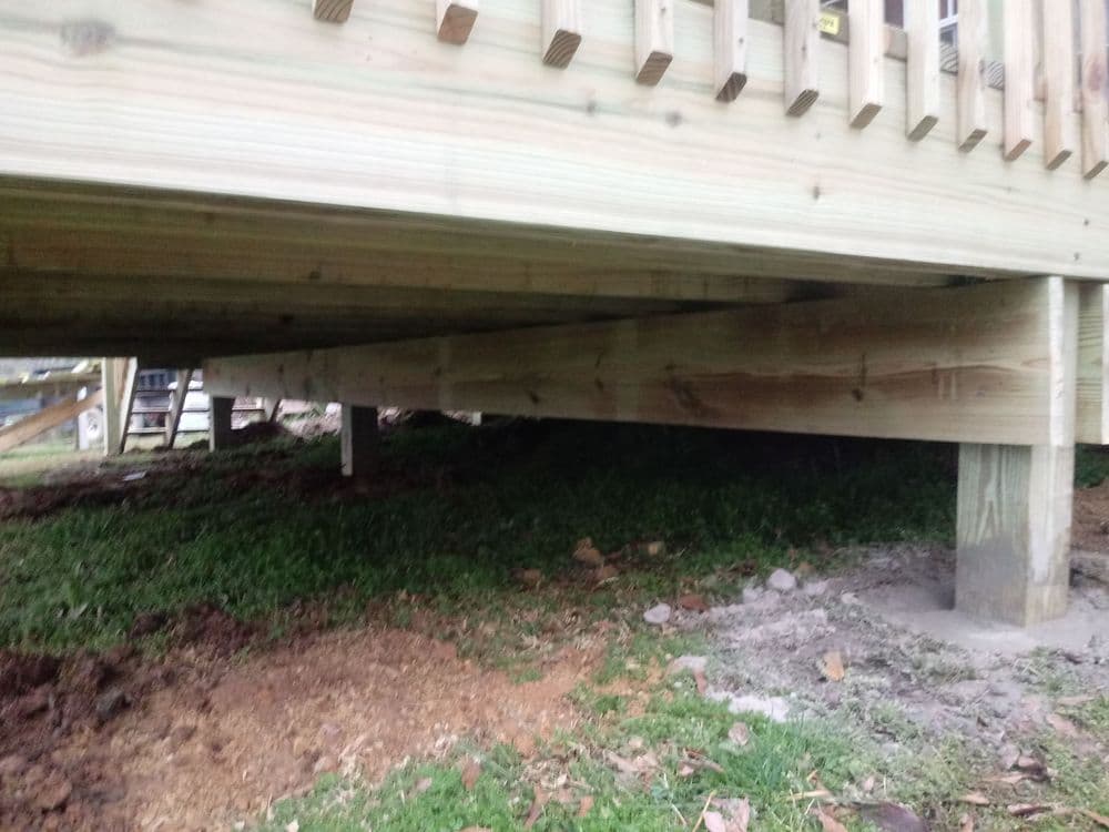 Underneath view of a wooden deck showing support beams and concrete footings on grass.