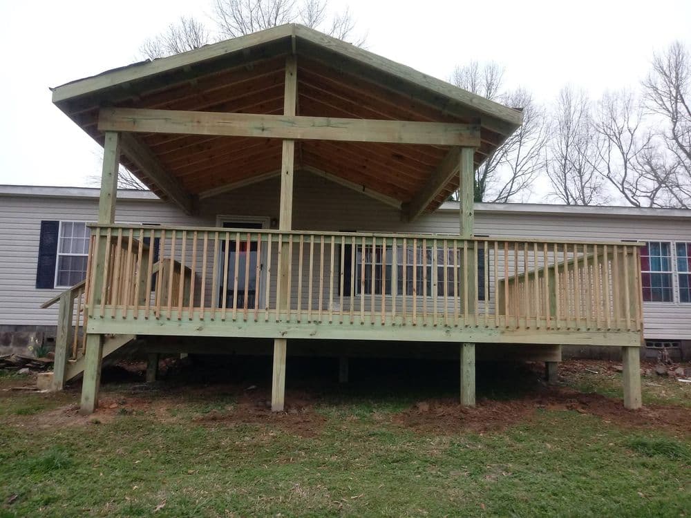 Wooden deck with a gabled roof, attached to a house, surrounded by greenery.