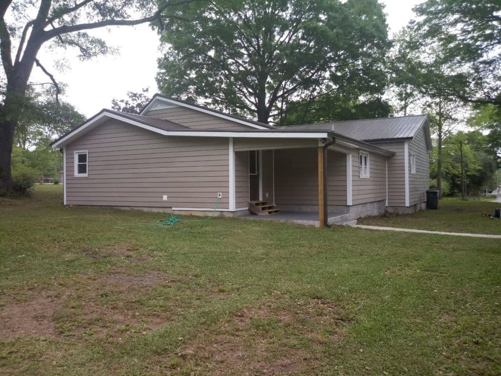 Single-story beige house with a porch, surrounded by green lawn and trees.