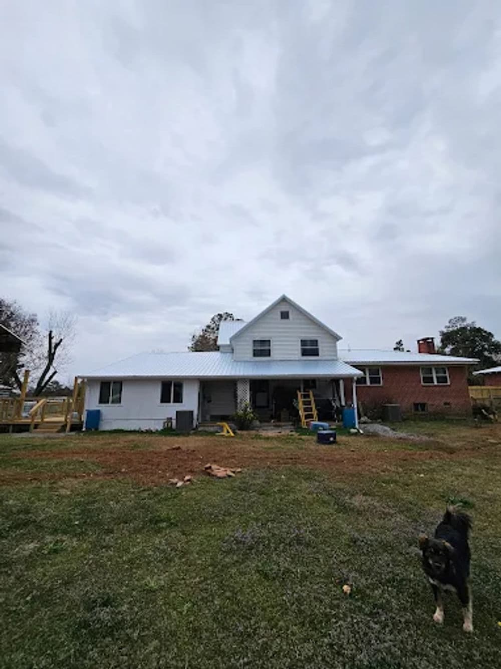 White farmhouse with gray roof on cloudy day, dog in foreground, construction area visible.